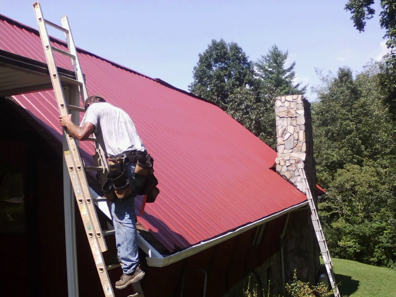 Roofer on a ladder beside a red metal roof and stone chimney — Soffit & Fascia Repair by Roofing Brothers in Onslow
